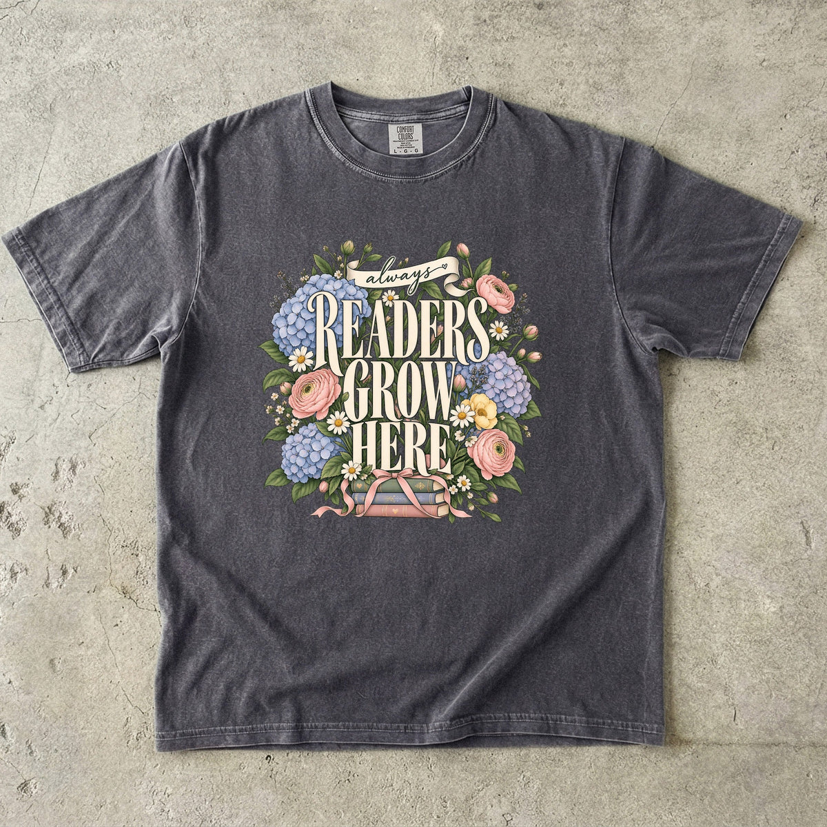 Pepper T-shirt featuring “always READERS GROW HERE” with large cream lettering surrounded by pastel blue flowers, pink blooms, daisies, leaves and ribbon-tied books.