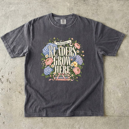 Pepper T-shirt featuring “always READERS GROW HERE” with large cream lettering surrounded by pastel blue flowers, pink blooms, daisies, leaves and ribbon-tied books.