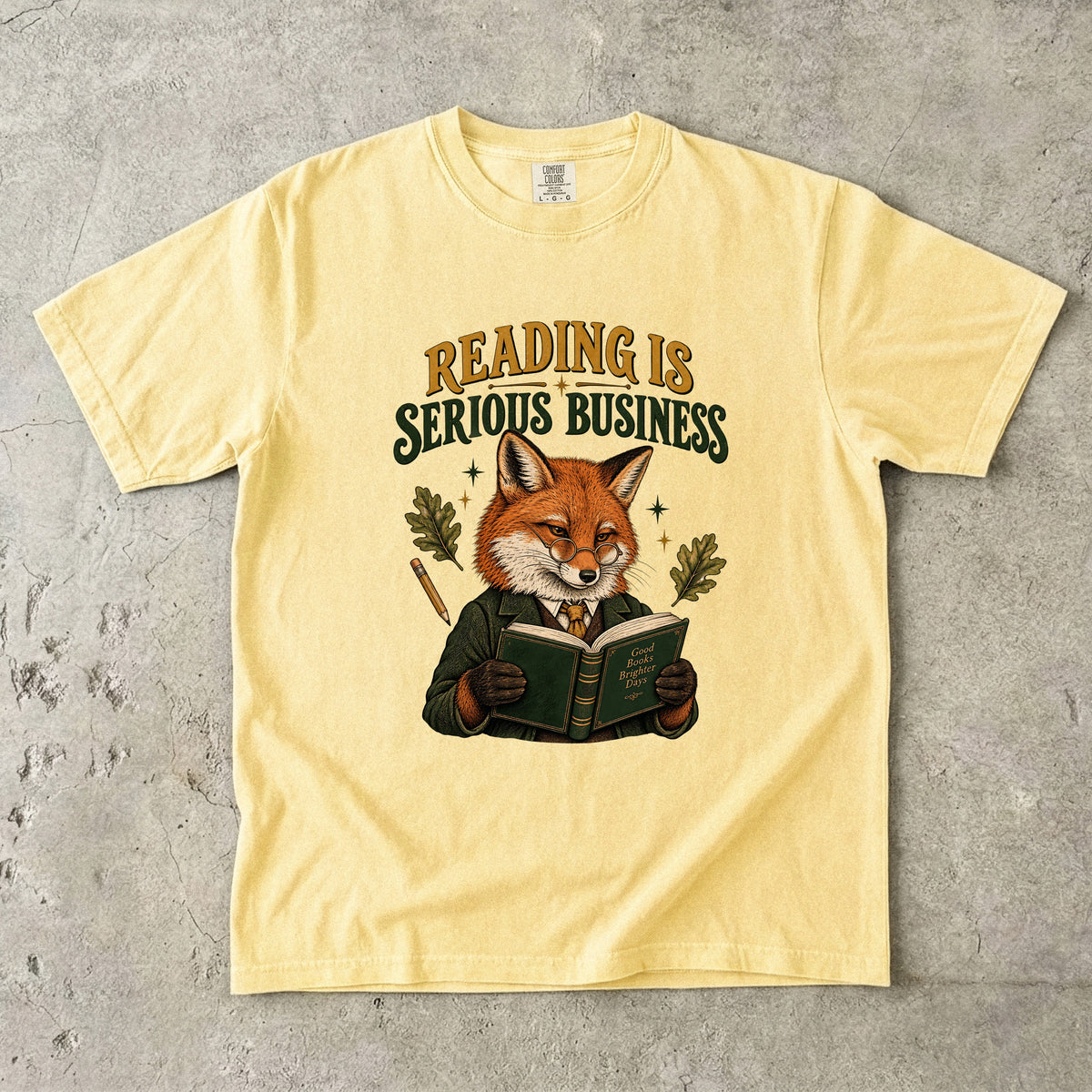 Butter T-shirt featuring “Reading Is Serious Business” with a fox in glasses and a green jacket reading an open book, with a pencil, oak leaves and stars.