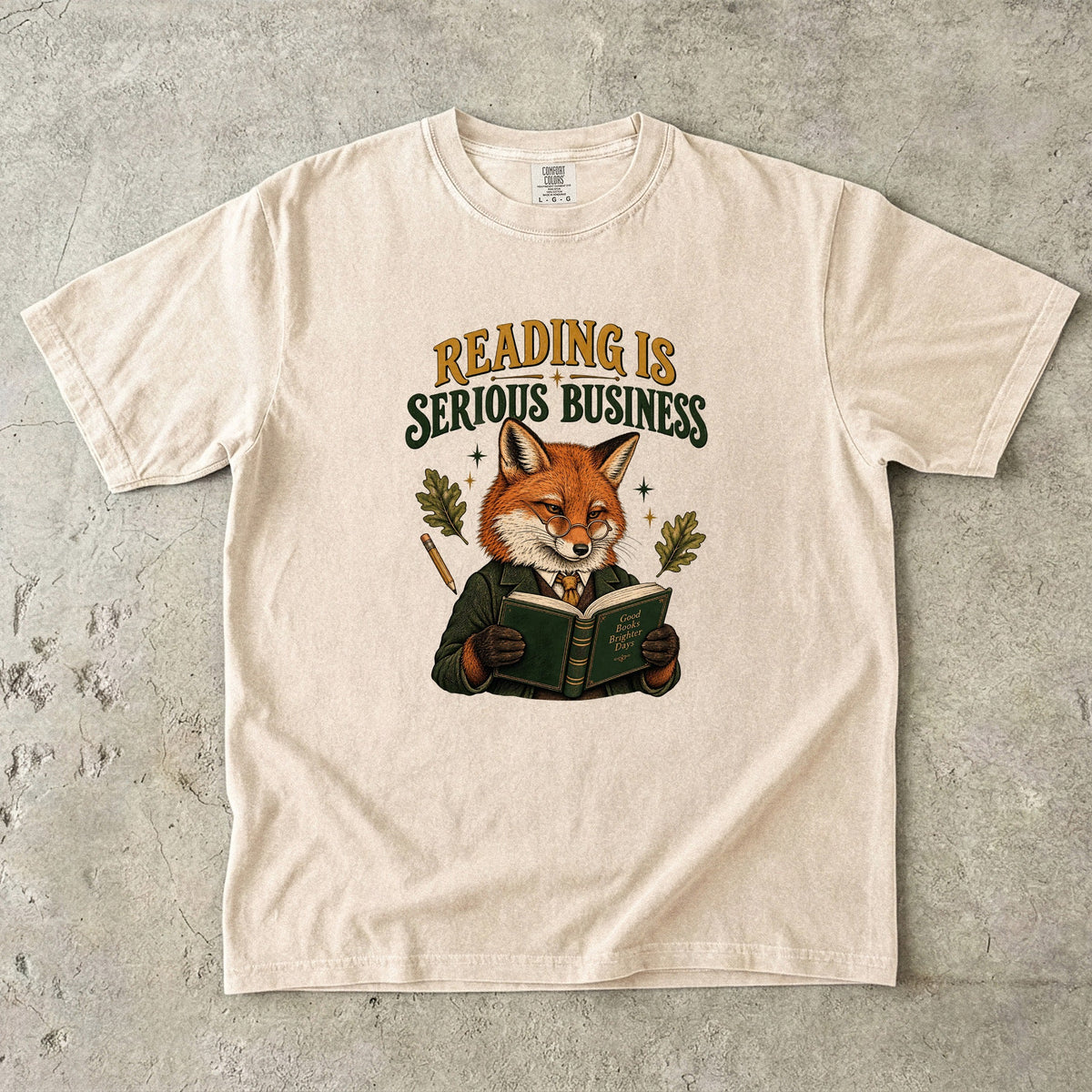 Ivory T-shirt featuring “Reading Is Serious Business” with a fox in glasses and a green jacket reading an open book, with a pencil, oak leaves and stars.
