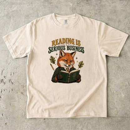 Ivory T-shirt featuring “Reading Is Serious Business” with a fox in glasses and a green jacket reading an open book, with a pencil, oak leaves and stars.