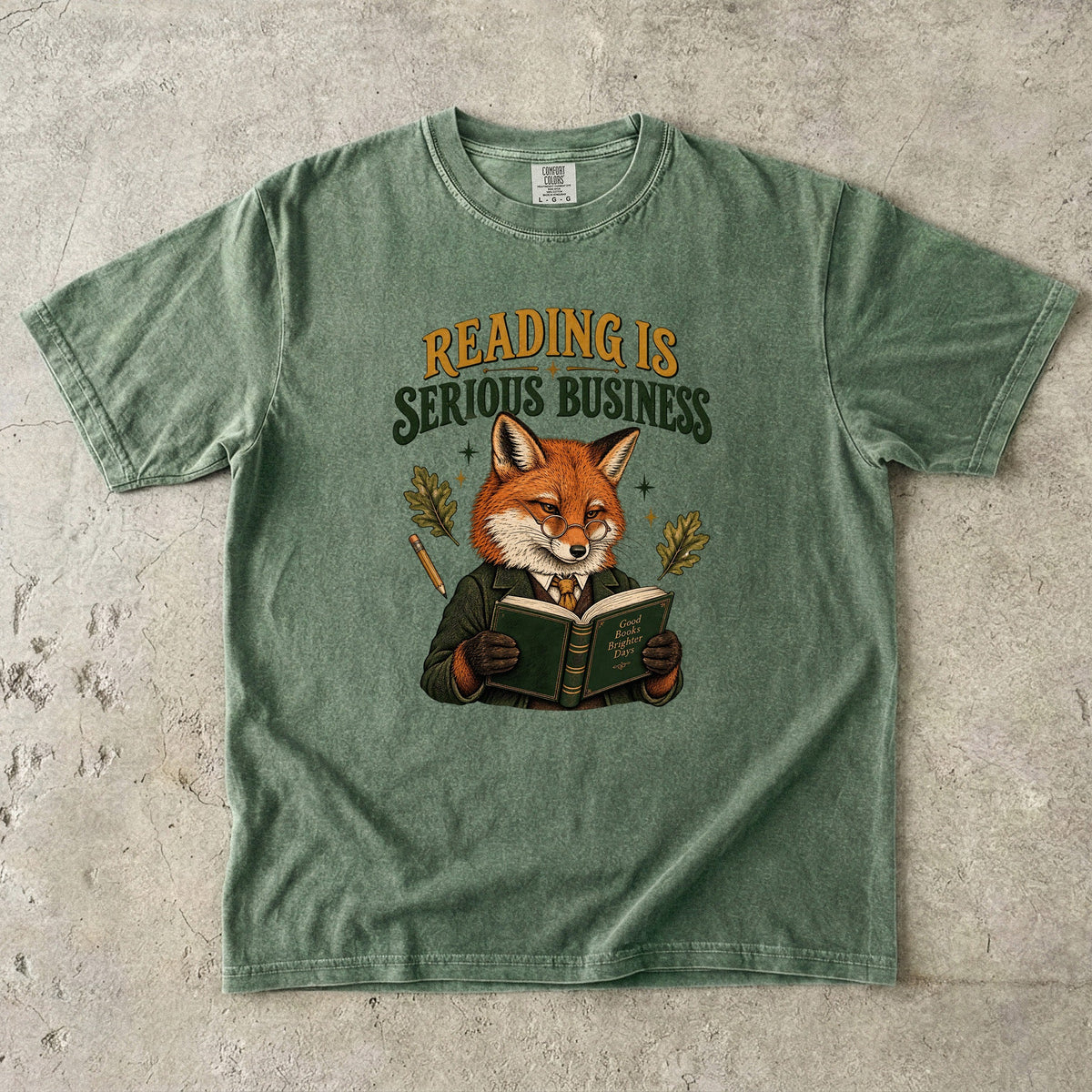 Moss T-shirt featuring “Reading Is Serious Business” with a fox in glasses and a green jacket reading an open book, with a pencil, oak leaves and stars.