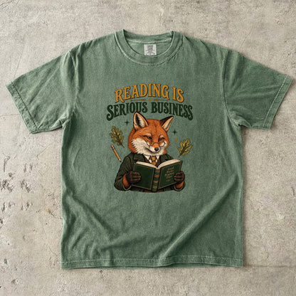 Moss T-shirt featuring “Reading Is Serious Business” with a fox in glasses and a green jacket reading an open book, with a pencil, oak leaves and stars.