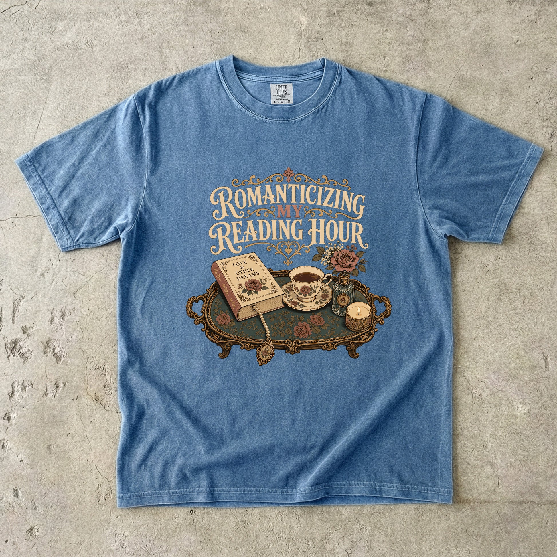 Front view of a Blue Jean Comfort Colors® book lover T-shirt featuring the Romanticizing My Reading Hour vintage design with a romance novel, teacup, candle, roses and ornate reading tray.