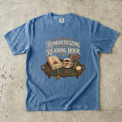 Front view of a Blue Jean Comfort Colors® book lover T-shirt featuring the Romanticizing My Reading Hour vintage design with a romance novel, teacup, candle, roses and ornate reading tray.