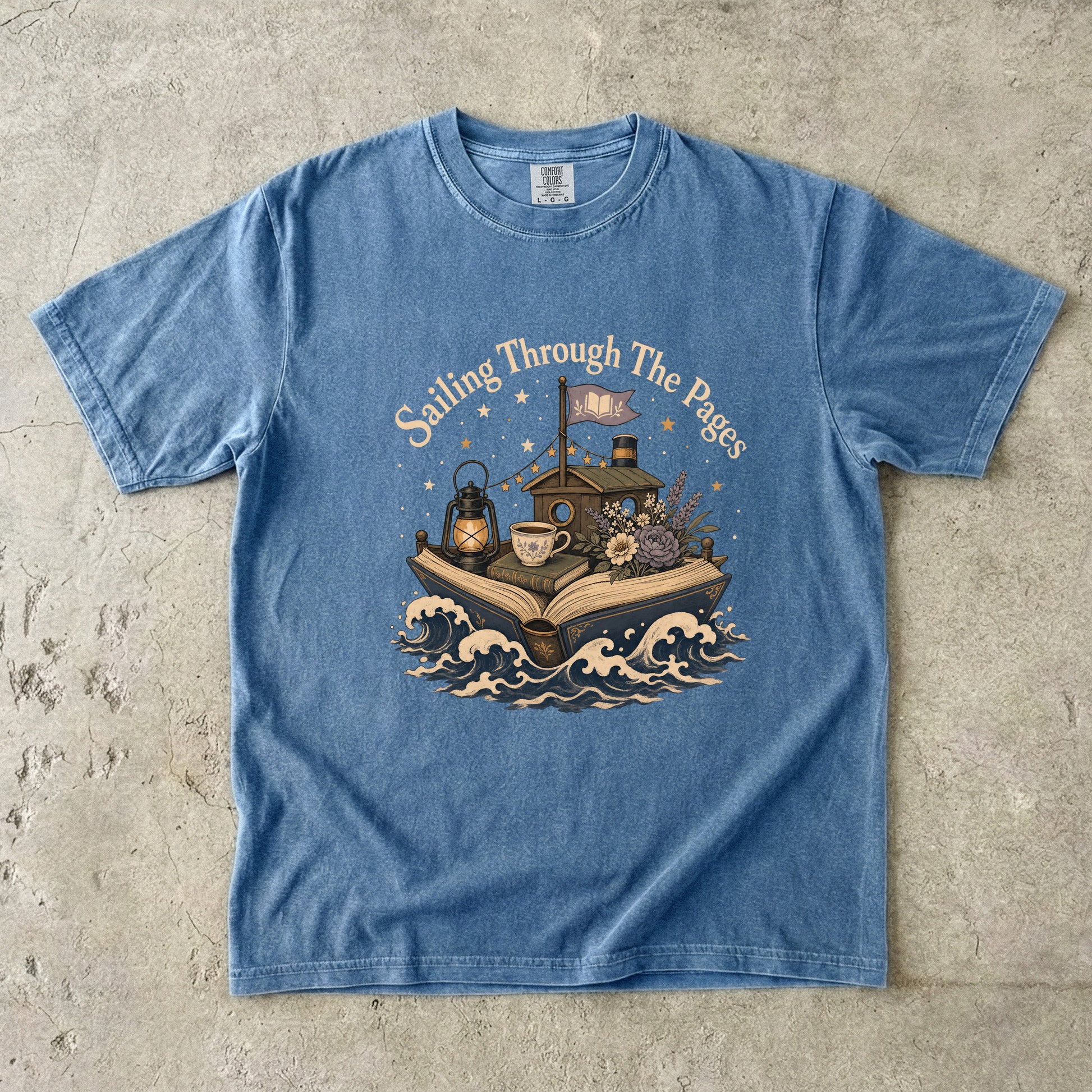 Front view of a Blue Jean Comfort Colors Book Lover T-shirt featuring the Sailing Through the Pages illustration with an open book boat, lantern, teacup and flowers.