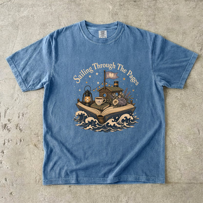 Front view of a Blue Jean Comfort Colors Book Lover T-shirt featuring the Sailing Through the Pages illustration with an open book boat, lantern, teacup and flowers.