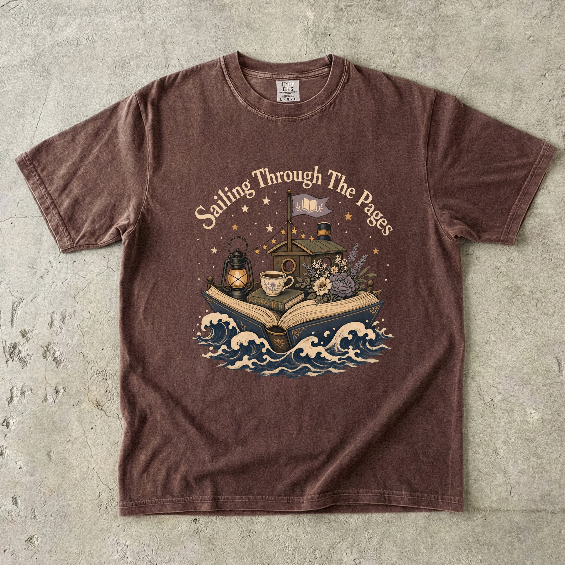 Front view of a Expresso Comfort Colors® Book Lover T-shirt featuring the "Sailing Through the Pages" vintage literary illustration with an open book boat, waves, lantern and cozy cottage.