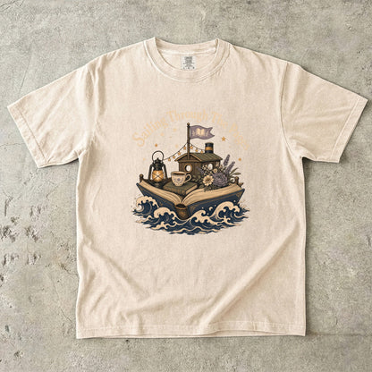 Front view of a Ivory  Comfort Colors Book Lover T-shirt featuring the Sailing Through the Pages illustration with an open book boat, lantern, teacup and flowers.