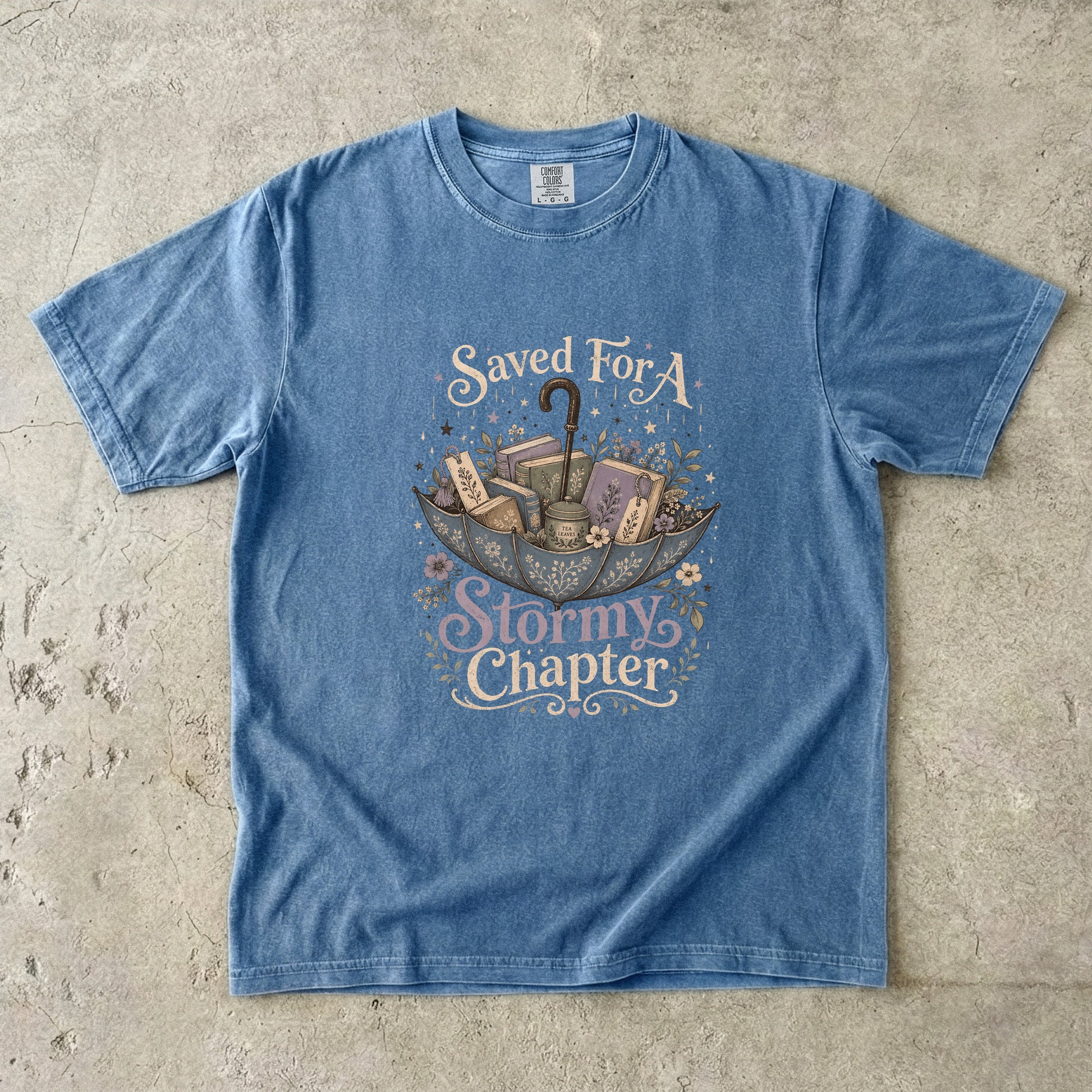 Saved for a Stormy Chapter reading T-shirt in Blue Jean featuring an umbrella filled with books, tea, bookmarks and flowers