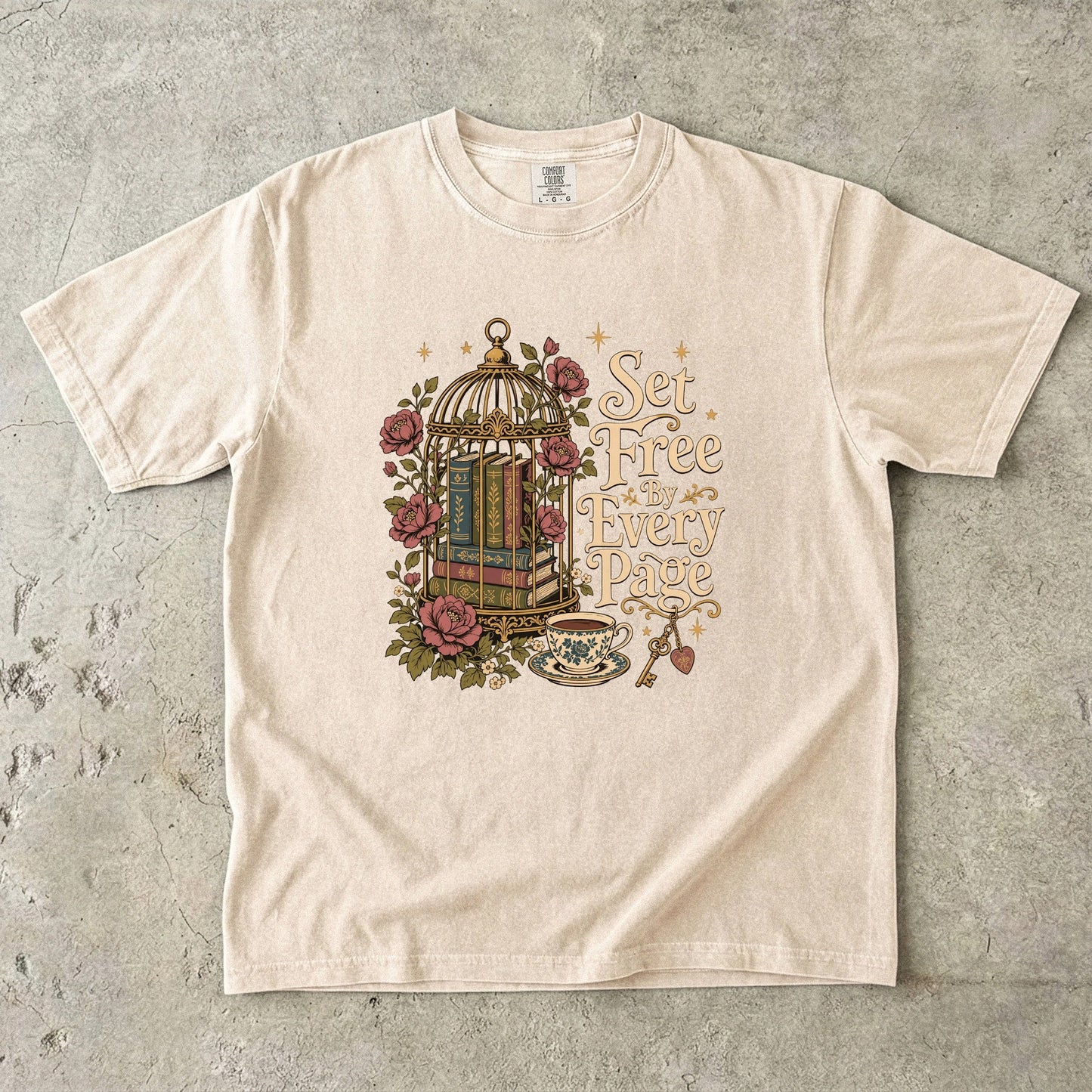 Flat lay of an Ivory Comfort Colors book lover T-shirt featuring the "Set Free By Every Page" vintage birdcage illustration with antique books, roses, teacup and ornate key.
