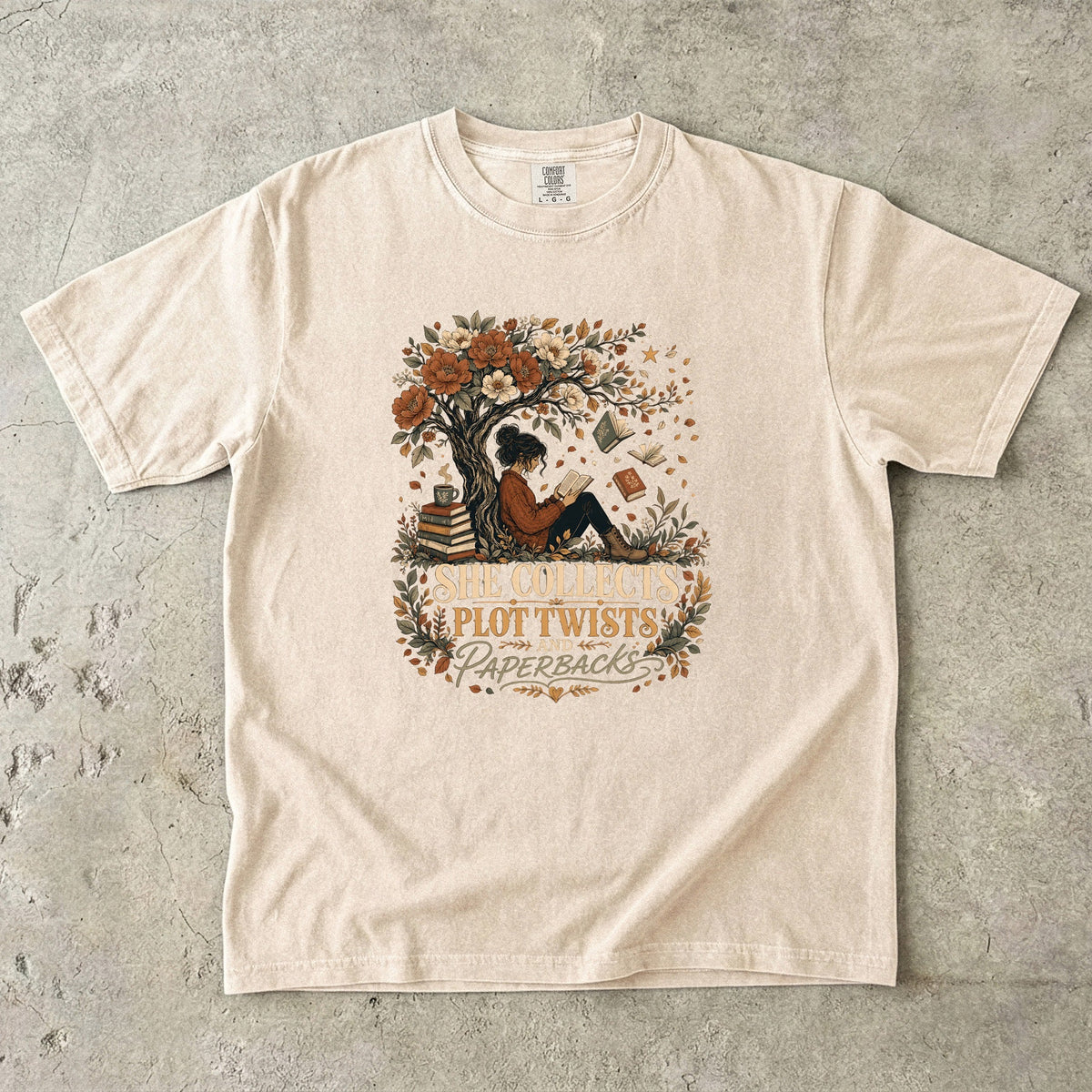 Ivory reading T-shirt featuring She Collects Plot Twists and Paperbacks with a reader beneath a flowering tree surrounded by books.