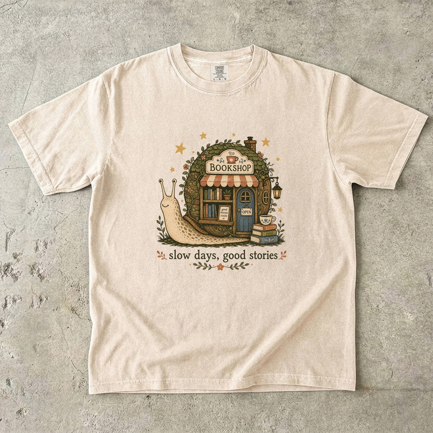 Slow Days, Good Stories bookshop T-shirt in Ivory featuring a snail beside a tiny bookshop with books and tea