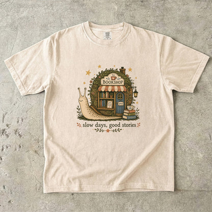 Slow Days, Good Stories bookshop T-shirt in Ivory featuring a snail beside a tiny bookshop with books and tea