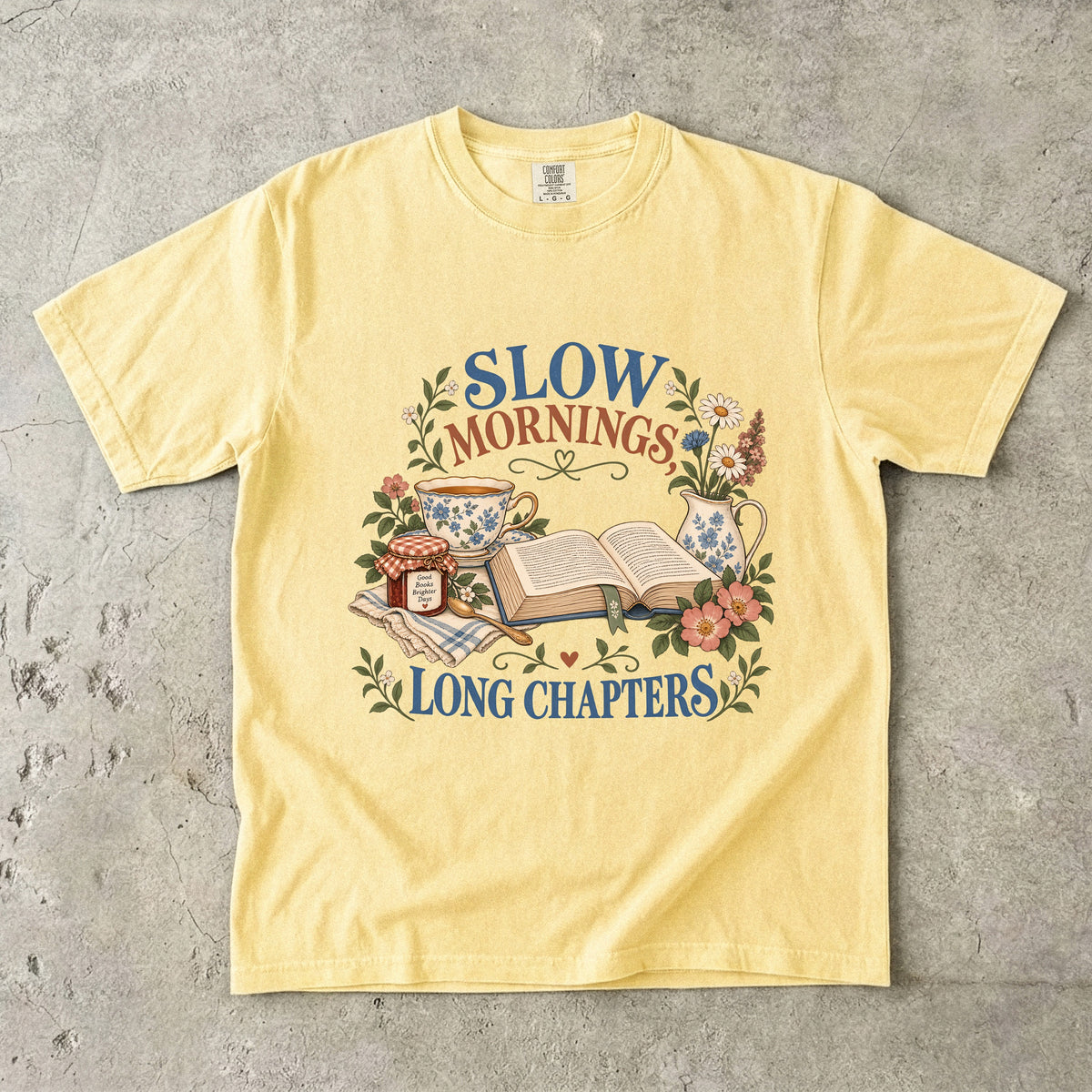 Butter T-shirt featuring “Slow Mornings, Long Chapters” with an open book, blue floral teacup and pitcher, a jam jar, spoon, flowers and checked cloth.