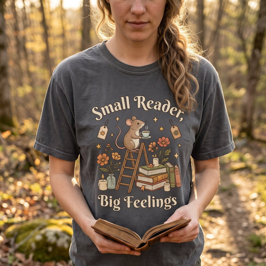 Lifestyle image of a model wearing the Comfort Colors 1717 T-shirt in Pepper featuring the "Small Reader, Big Feelings" vintage reading mouse illustration while holding an open book on a woodland path.

