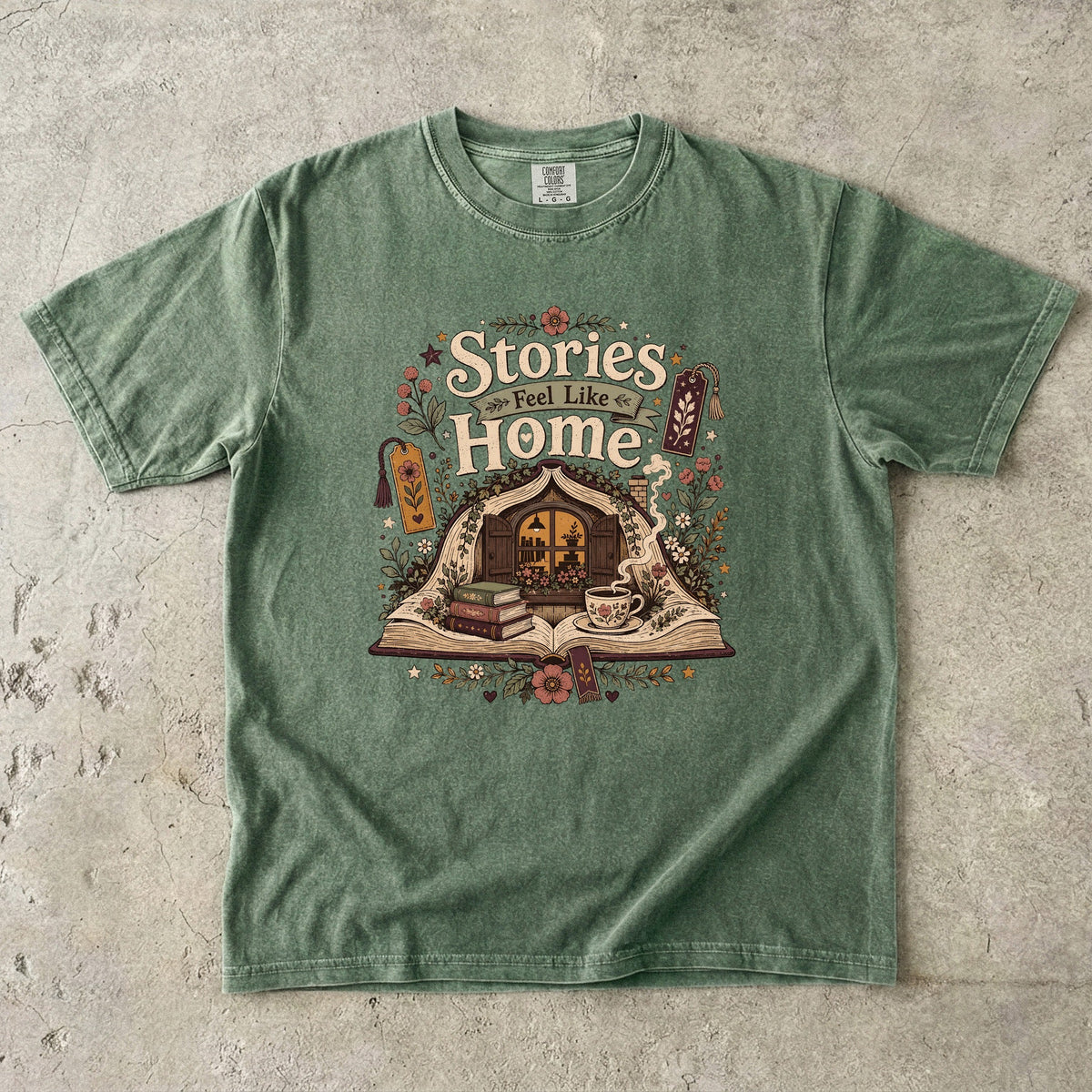 Stories Feel Like Home book lover T-shirt with open book, cozy reading nook, stacked books, teacup and floral details on Moss.