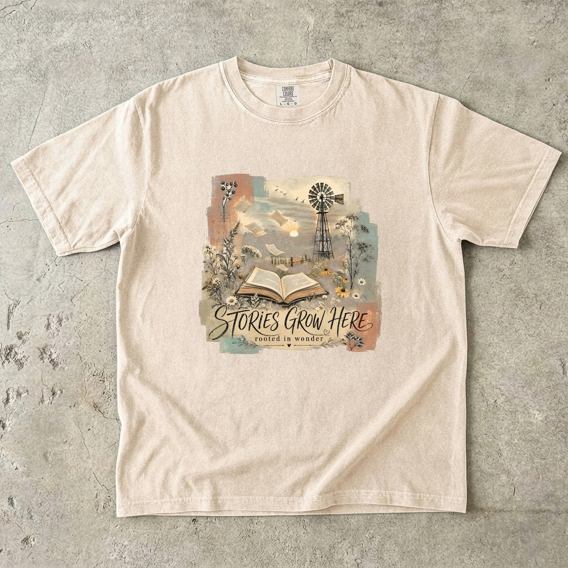 Ivory Comfort Colors Stories Grow Here T-shirt with an open book, wildflowers, windmill and dreamy landscape.