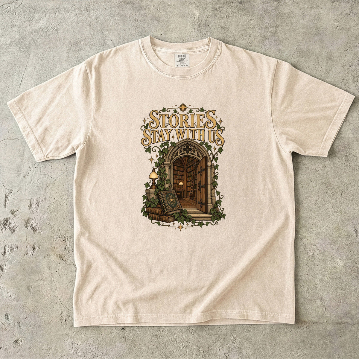 Ivory T-shirt featuring “Stories Stay With Us” with an ivy-covered arched library doorway, antique books, lamps and gold stars.