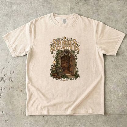 Ivory T-shirt featuring “Stories Stay With Us” with an ivy-covered arched library doorway, antique books, lamps and gold stars.