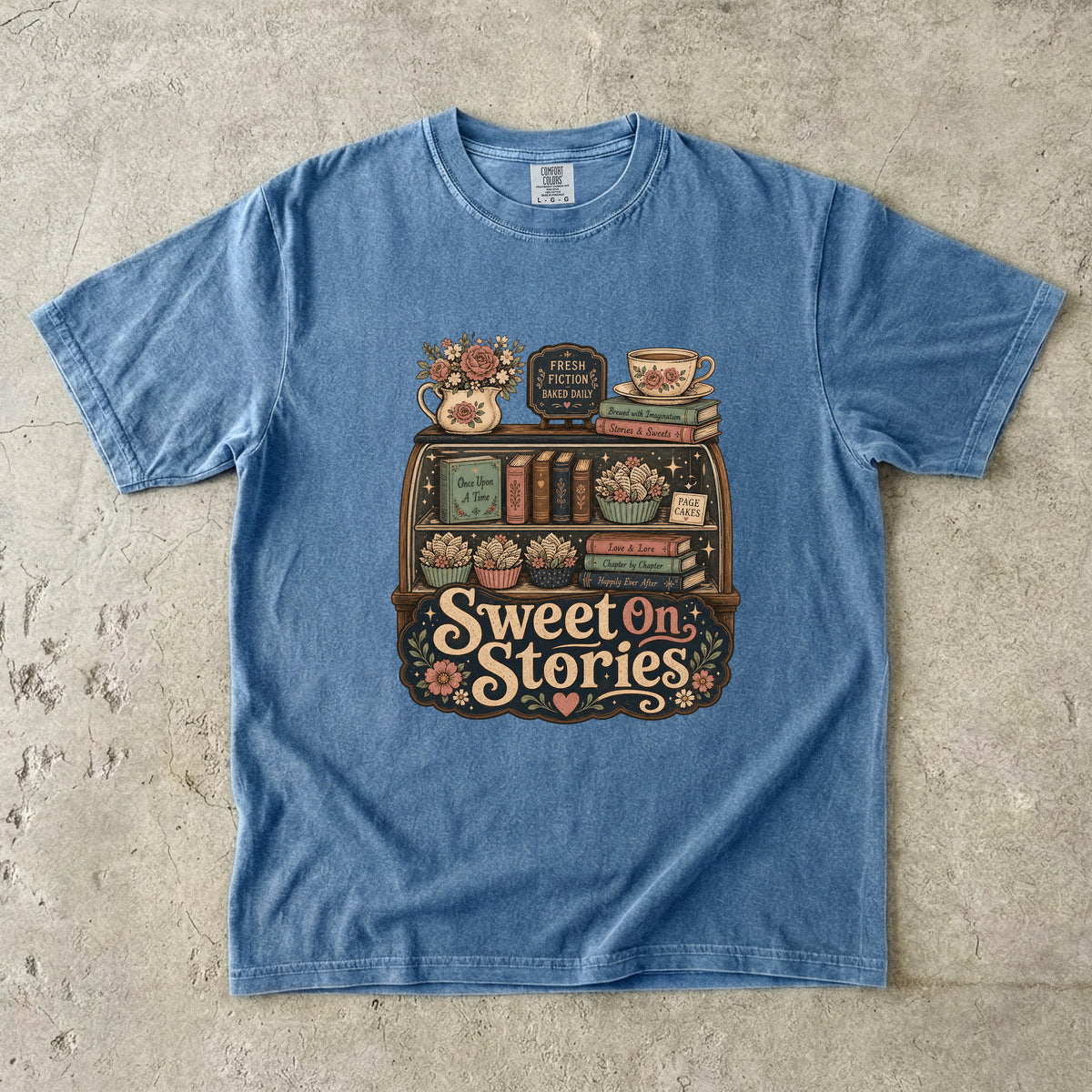 Sweet on Stories book lover T-shirt in Blue Jean featuring a vintage fresh-fiction display with books, page cakes, flowers and teacup