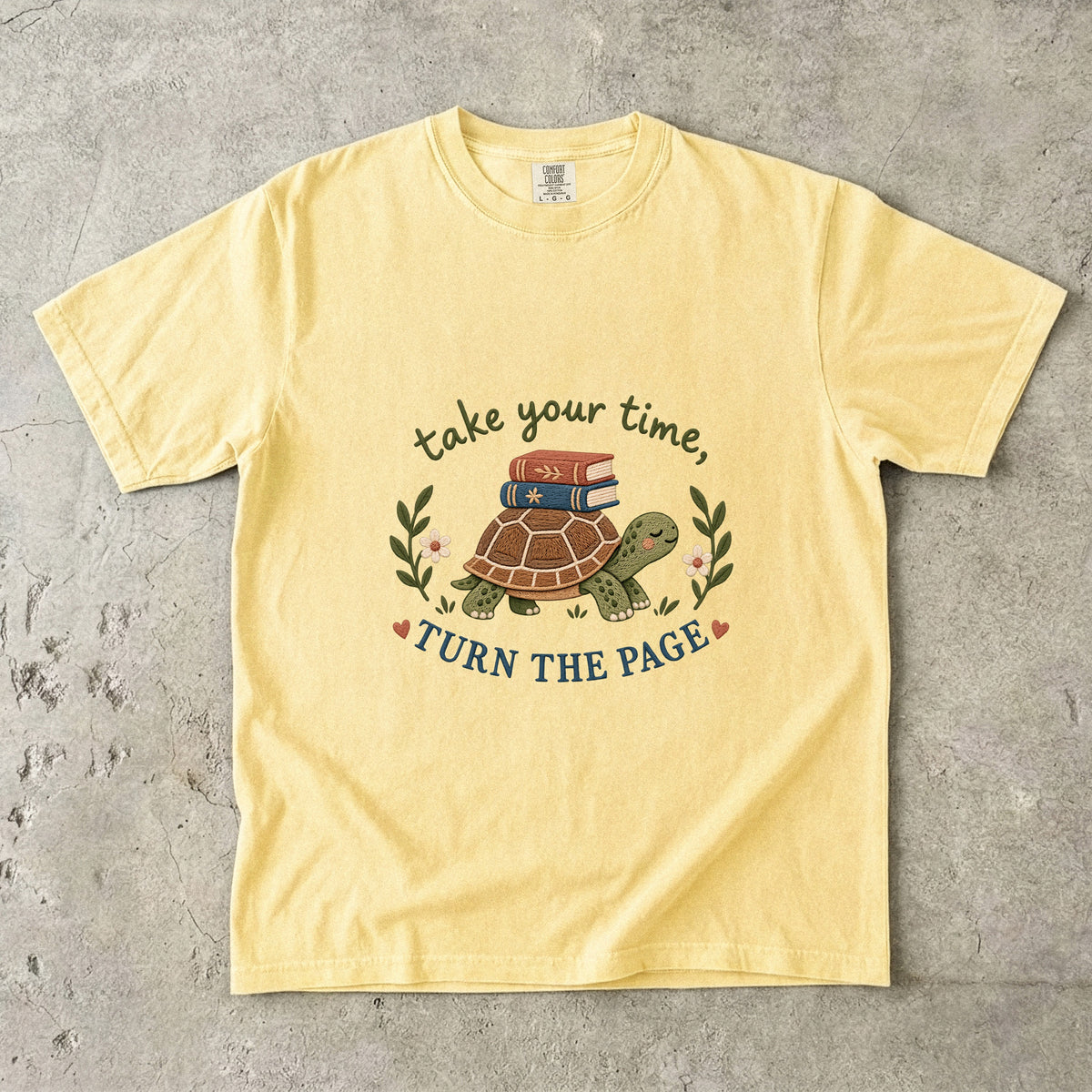 Butter T-shirt featuring “take your time, TURN THE PAGE” with a smiling turtle carrying two books, leafy sprigs, daisies and small hearts.