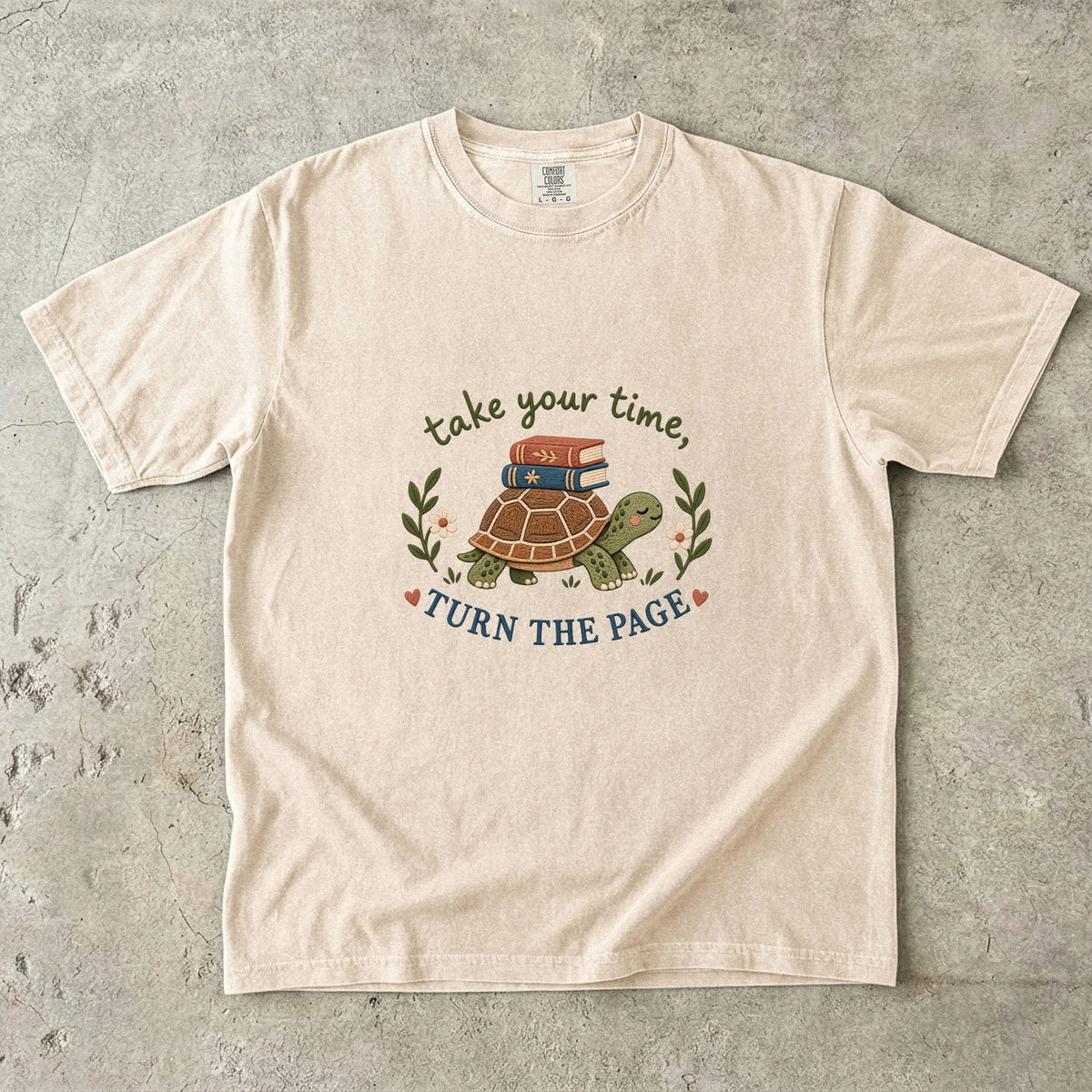 Ivory T-shirt featuring “take your time, TURN THE PAGE” with a smiling turtle carrying two books, leafy sprigs, daisies and small hearts.