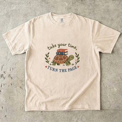 Ivory T-shirt featuring “take your time, TURN THE PAGE” with a smiling turtle carrying two books, leafy sprigs, daisies and small hearts.