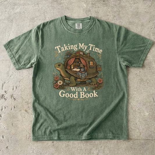 Front view of a Moss Comfort Colors® book lover T-shirt featuring the Taking My Time With a Good Book design with a turtle carrying a cosy reading nook, books and flowers.