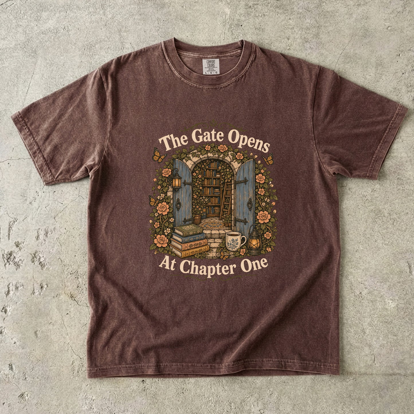 Front view of an Espresso Comfort Colors® Book Lover T-shirt featuring The Gate Opens at Chapter One hidden library illustration with vintage books, flowers, lanterns and open blue doors.