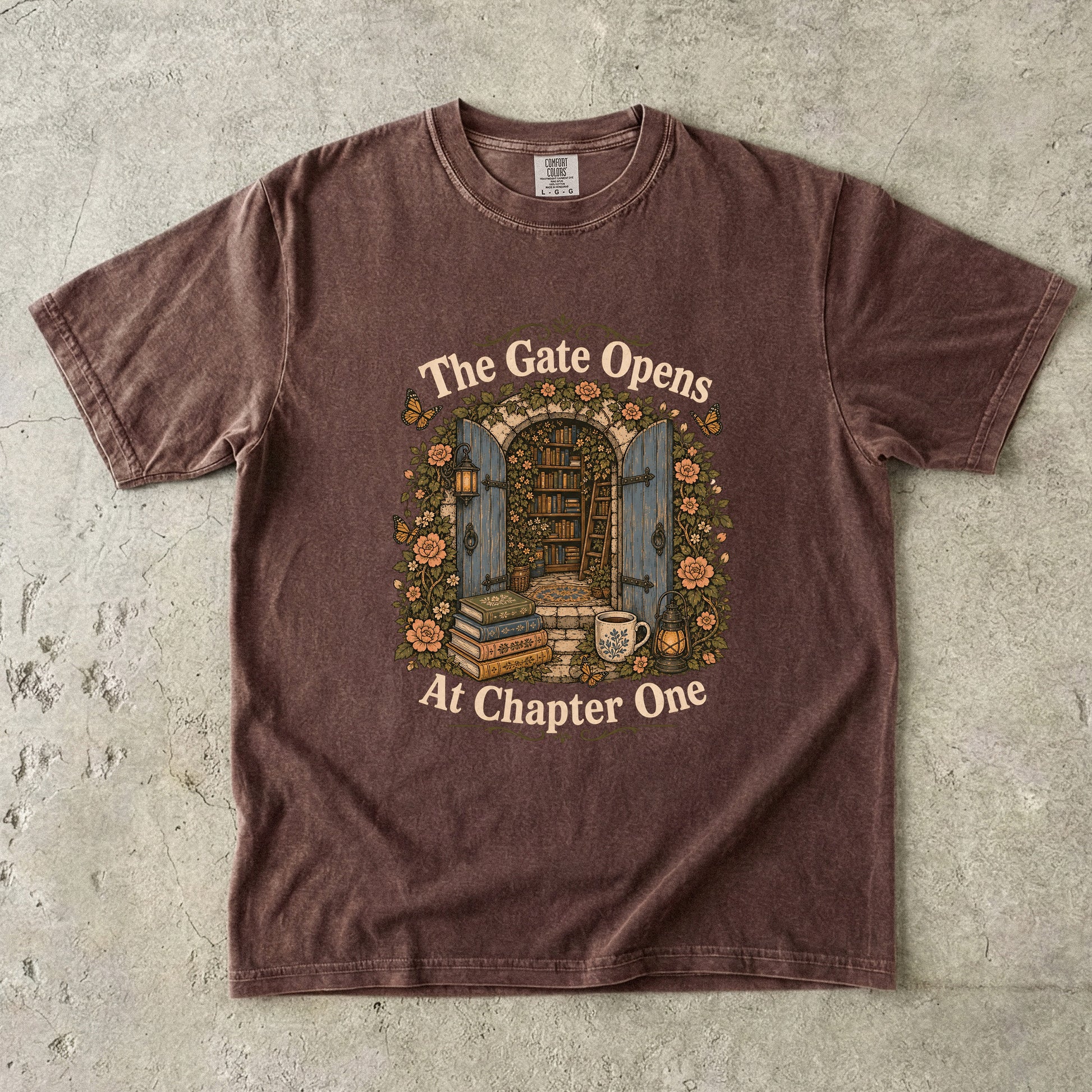 Front view of an Espresso Comfort Colors® Book Lover T-shirt featuring The Gate Opens at Chapter One hidden library illustration with vintage books, flowers, lanterns and open blue doors.