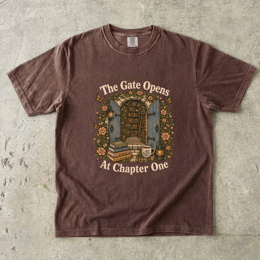 Front view of an Espresso Comfort Colors® Book Lover T-shirt featuring The Gate Opens at Chapter One hidden library illustration with vintage books, flowers, lanterns and open blue doors.