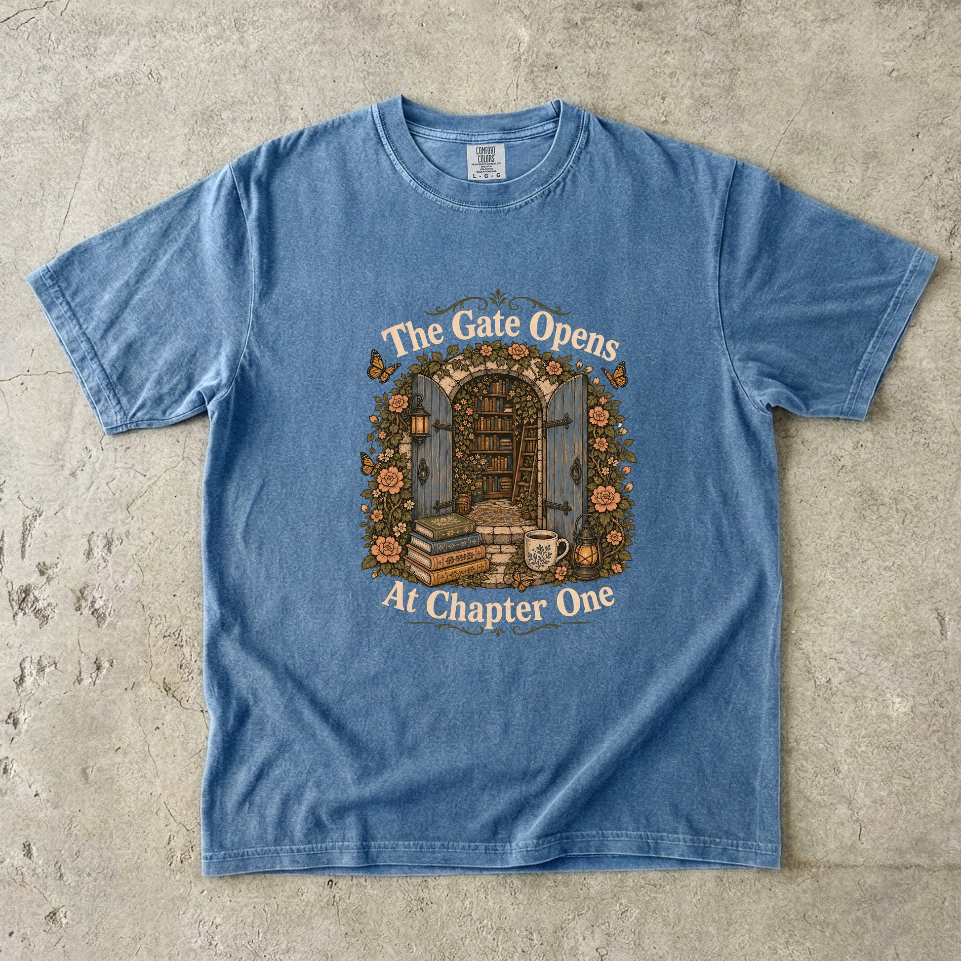 The Gate Opens at Chapter One literary T-shirt in Blue Jean featuring open gates leading into a flower-filled hidden library