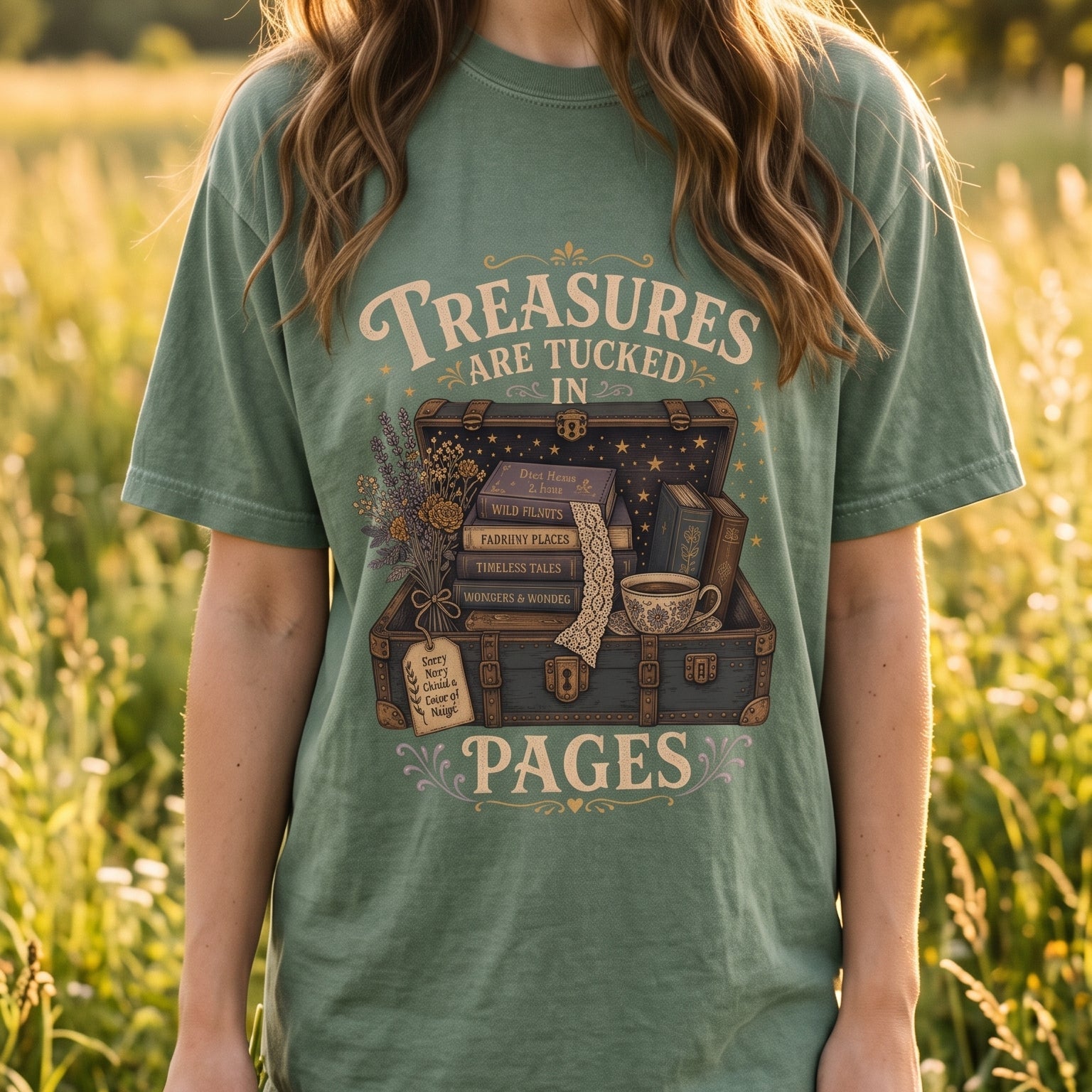 Lifestyle image of a model wearing the Comfort Colors 1717 T-shirt in Seafoam featuring the "Treasures Are Tucked in Pages" vintage illustration with an open treasure chest, books, flowers and a teacup in a wildflower meadow. 