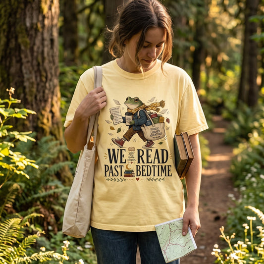 Reader wearing a We Read Past Bedtime book lover T-shirt while carrying books and a tote on a woodland path.