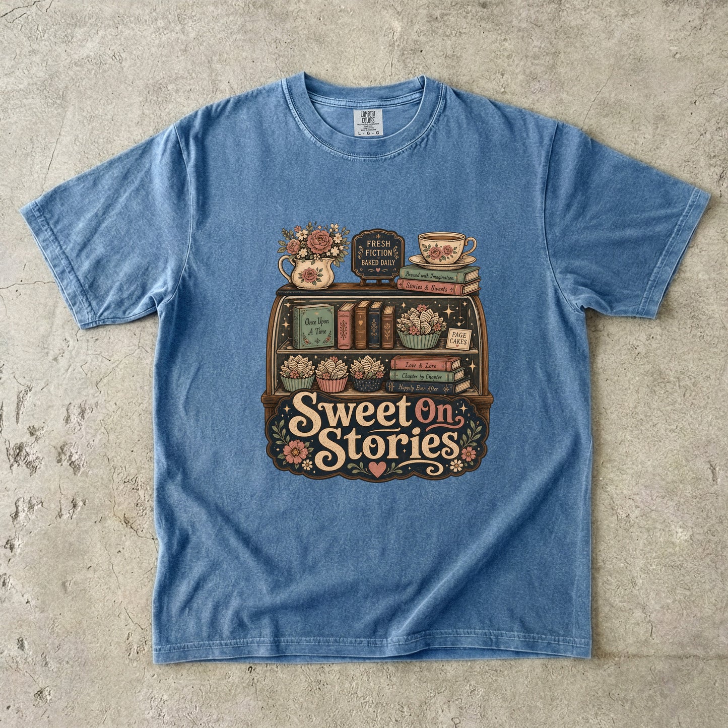 Front view of a Blue Jean Comfort Colors® book lover T-shirt featuring the Sweet On Stories vintage bookstore display with books, flowers and tea-inspired artwork.